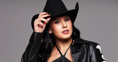 A cantora Ana Castela durante apresentação, vestindo chapéu e look country, sorrindo e segurando o microfone no palco iluminado.