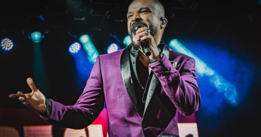 Imagem de Alexandre Pires iluminado no palco, microfone em mãos, clima festivo e luzes quentes ao fundo, transmitindo energia e emoção do show Pagonejo Bão.