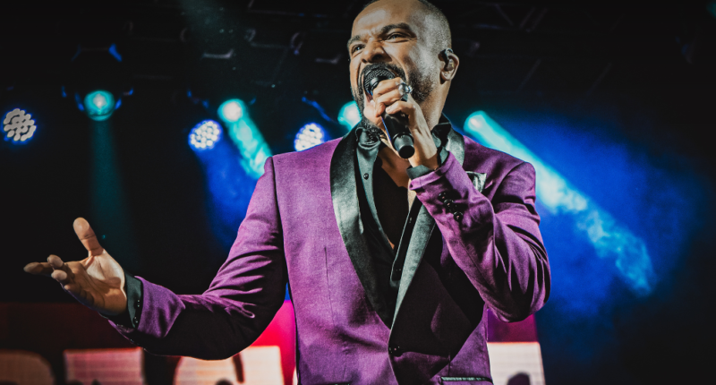 Imagem de Alexandre Pires iluminado no palco, microfone em mãos, clima festivo e luzes quentes ao fundo, transmitindo energia e emoção do show Pagonejo Bão.