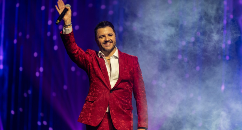 Maurício Manieri no palco durante o espetáculo Classics Super Hits, com iluminação cênica e banda ao fundo.