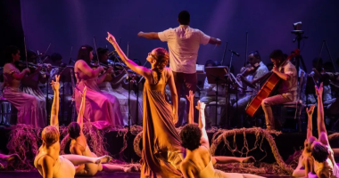 Bailarinos e músicos em cena com iluminação cênica representando o Pantanal em espetáculo artístico