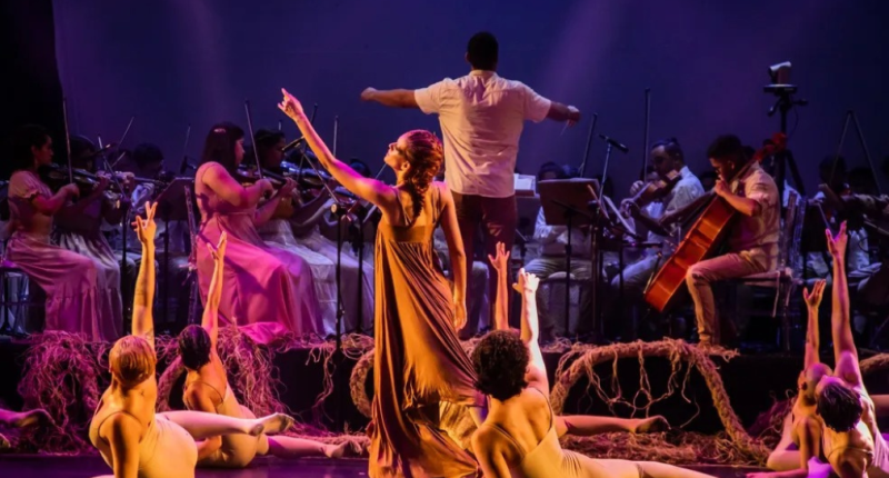 Bailarinos e músicos em cena com iluminação cênica representando o Pantanal em espetáculo artístico