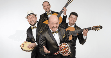 Grupo Demônios da Garoa se apresenta em palco iluminado, com cenário intimista e painel de LED ao fundo