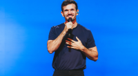 Padre Patrick em palco iluminado, interagindo com o público em espetáculo de humor e reflexão