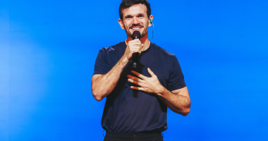 Padre Patrick em palco iluminado, interagindo com o público em espetáculo de humor e reflexão