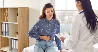 mulher sentada sozinha em ambiente fechado segurando a barriga em expressão de dor e desconforto intestinal