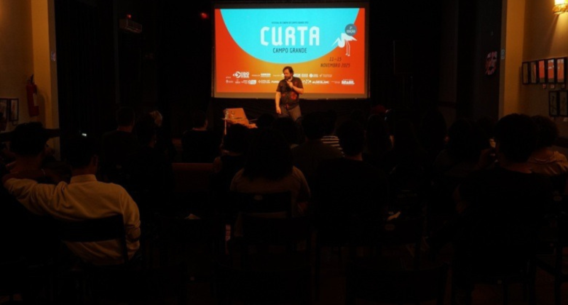diretor conversa com público durante festival de cinema em auditório com projeção de curta-metragem