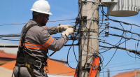 Profissional eletricista realiza manutenção em rede elétrica urbana utilizando equipamentos de segurança, junto a poste com fiação e transformador, durante atividade de campo em dia ensolarado.