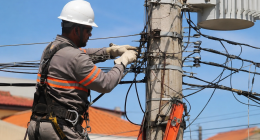 Profissional eletricista realiza manutenção em rede elétrica urbana utilizando equipamentos de segurança, junto a poste com fiação e transformador, durante atividade de campo em dia ensolarado.