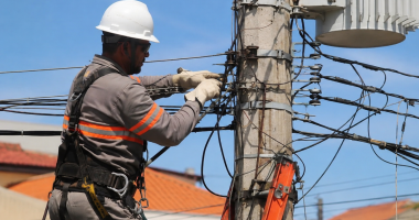 Profissional eletricista realiza manutenção em rede elétrica urbana utilizando equipamentos de segurança, junto a poste com fiação e transformador, durante atividade de campo em dia ensolarado.