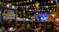 Famílias circulam entre barracas típicas durante festa junina comunitária, enquanto apresentações culturais acontecem em palco ao ar livre.