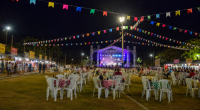 Vista ampla de uma festa junina realizada em um campo de futebol durante a noite, com bandeirinhas coloridas decorando todo o espaço. Ao centro, um palco iluminado recebe apresentações musicais, enquanto dezenas de mesas com toalhas xadrez e cadeiras plásticas ocupam o gramado. Nas laterais, barracas de comidas típicas atendem o público, criando um cenário tradicional de celebração comunitária ao ar livre.