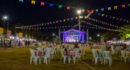 Vista ampla de uma festa junina realizada em um campo de futebol durante a noite, com bandeirinhas coloridas decorando todo o espaço. Ao centro, um palco iluminado recebe apresentações musicais, enquanto dezenas de mesas com toalhas xadrez e cadeiras plásticas ocupam o gramado. Nas laterais, barracas de comidas típicas atendem o público, criando um cenário tradicional de celebração comunitária ao ar livre.