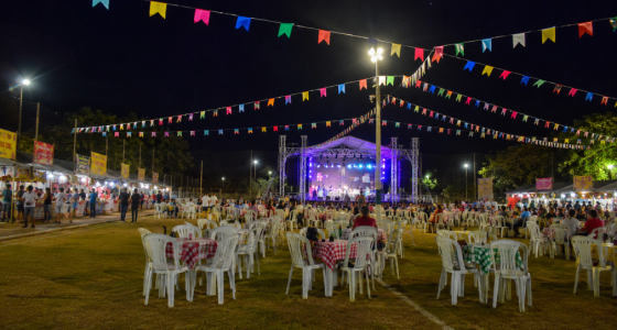 Vista ampla de uma festa junina realizada em um campo de futebol durante a noite, com bandeirinhas coloridas decorando todo o espaço. Ao centro, um palco iluminado recebe apresentações musicais, enquanto dezenas de mesas com toalhas xadrez e cadeiras plásticas ocupam o gramado. Nas laterais, barracas de comidas típicas atendem o público, criando um cenário tradicional de celebração comunitária ao ar livre.