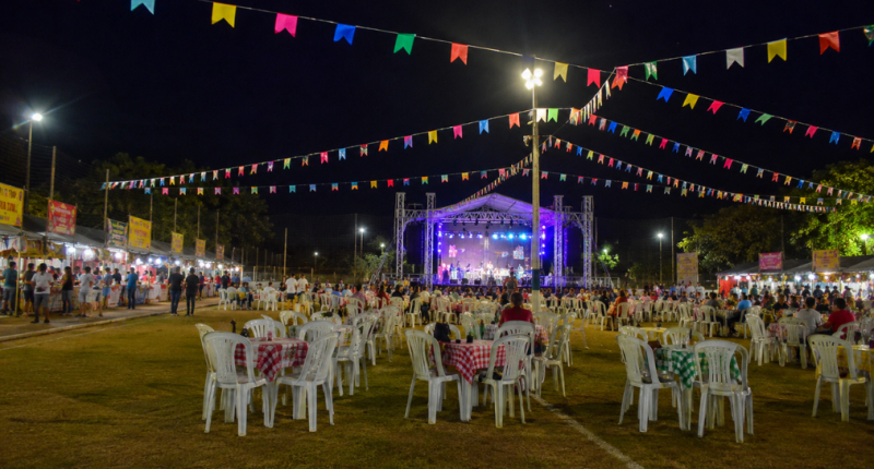 Vista ampla de uma festa junina realizada em um campo de futebol durante a noite, com bandeirinhas coloridas decorando todo o espaço. Ao centro, um palco iluminado recebe apresentações musicais, enquanto dezenas de mesas com toalhas xadrez e cadeiras plásticas ocupam o gramado. Nas laterais, barracas de comidas típicas atendem o público, criando um cenário tradicional de celebração comunitária ao ar livre.