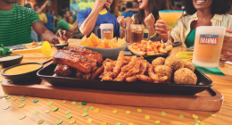 Grupo de amigos assiste a uma partida de futebol enquanto compartilha pratos do Outback, bebidas e momentos de confraternização em ambiente descontraído.