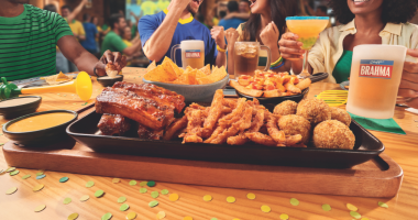 Grupo de amigos assiste a uma partida de futebol enquanto compartilha pratos do Outback, bebidas e momentos de confraternização em ambiente descontraído.