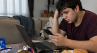 Jovem sentado diante do computador consome alimentos ultraprocessados enquanto utiliza o celular, ilustrando hábitos associados ao sedentarismo.