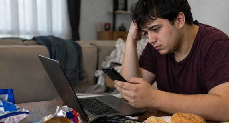 Jovem sentado diante do computador consome alimentos ultraprocessados enquanto utiliza o celular, ilustrando hábitos associados ao sedentarismo.