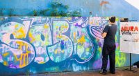 Artista pinta mural inspirado no sobá durante atividade do projeto Mural Postal Campão em Campo Grande.