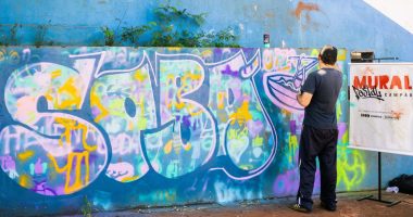 Artista pinta mural inspirado no sobá durante atividade do projeto Mural Postal Campão em Campo Grande.