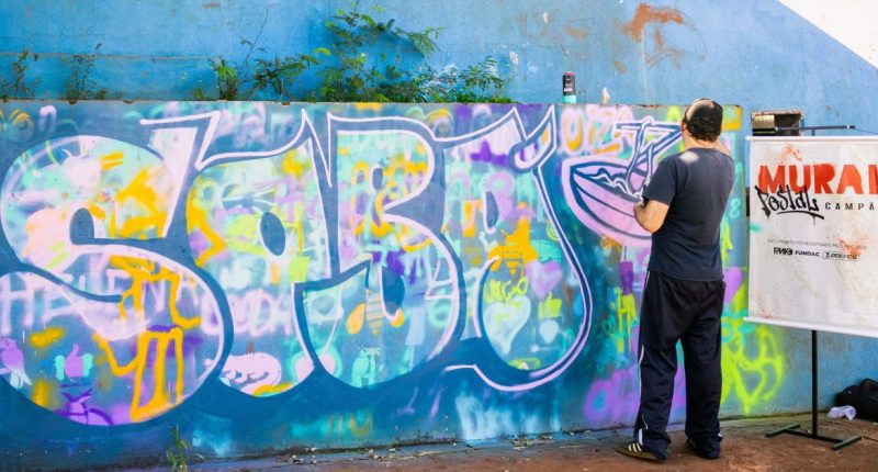 Artista pinta mural inspirado no sobá durante atividade do projeto Mural Postal Campão em Campo Grande.