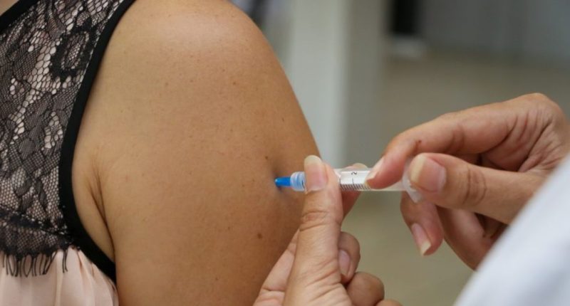 Profissional de saúde aplicando vacina no braço de adolescente em unidade de vacinação.